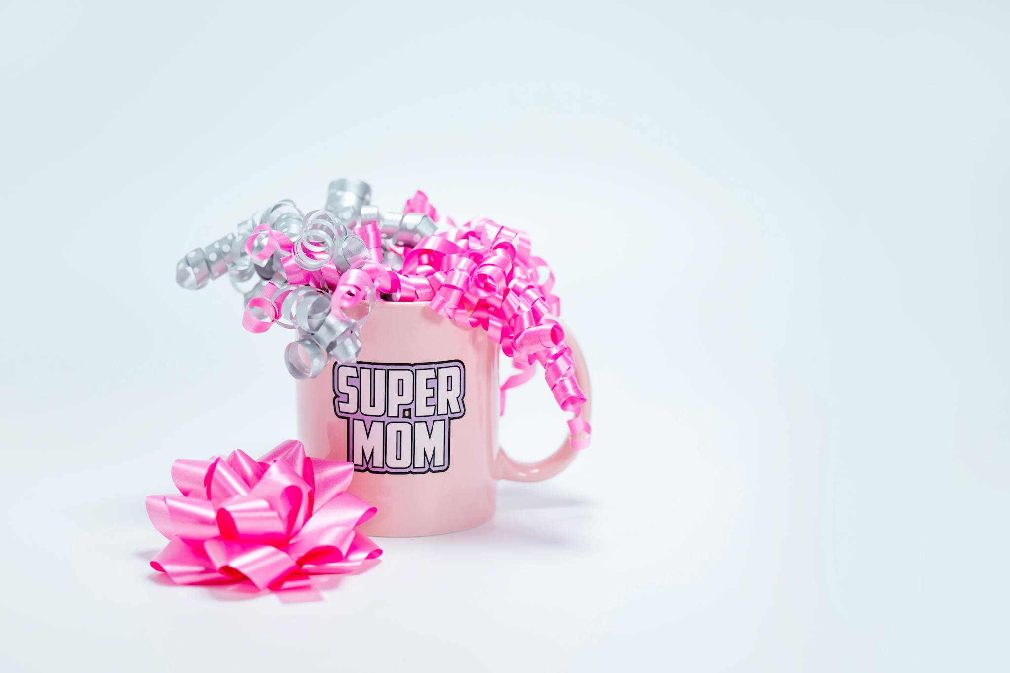 A pink Super Mom mug adorned with curly pink and silver ribbons on a white background, perfect for Mother's Day.