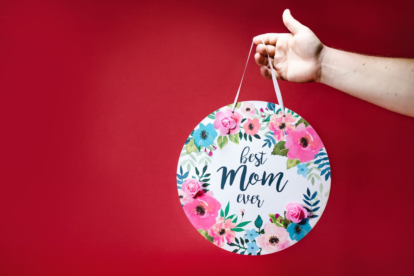 Round floral sign with 'Best Mom Ever' text held against a red background.